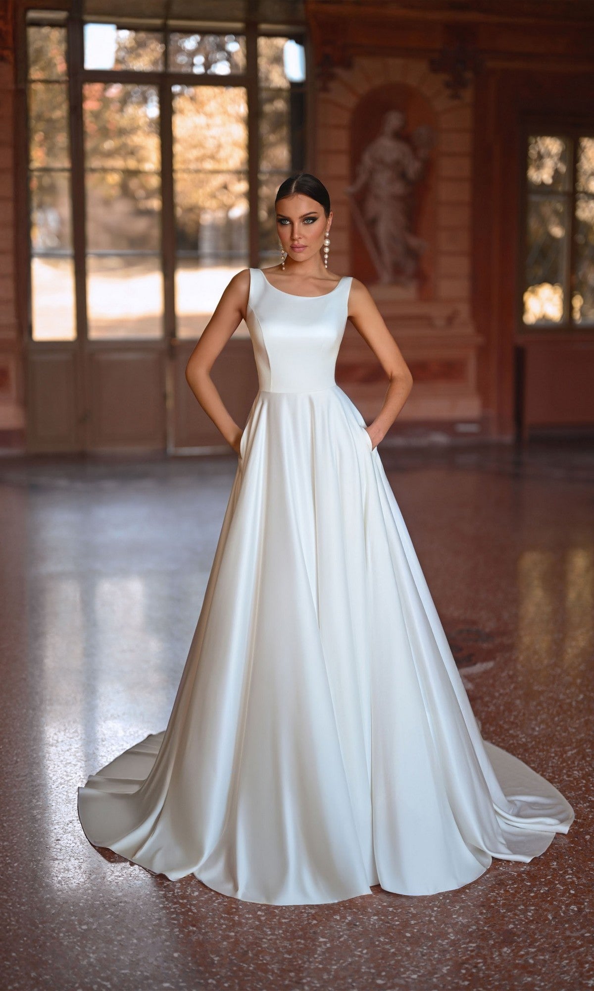 A woman wearing the Long White Dress: Alyce 7053, a sleeveless formal gown with an A-line skirt and pockets, stands in an ornate room with large windows and classical decor.